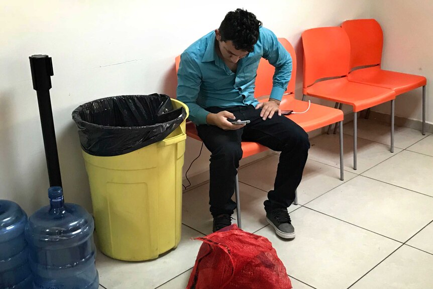 A deportee charges his phone after arriving at the reception centre, his belongings on the floor.
