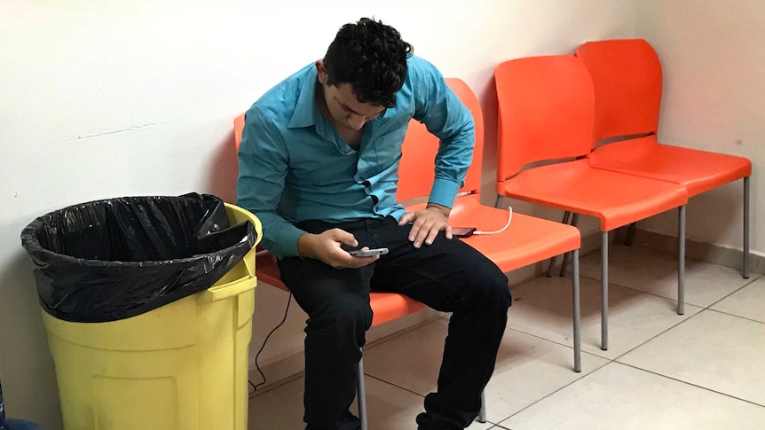 A deportee charges his phone after arriving at the reception centre, his belongings on the floor.
