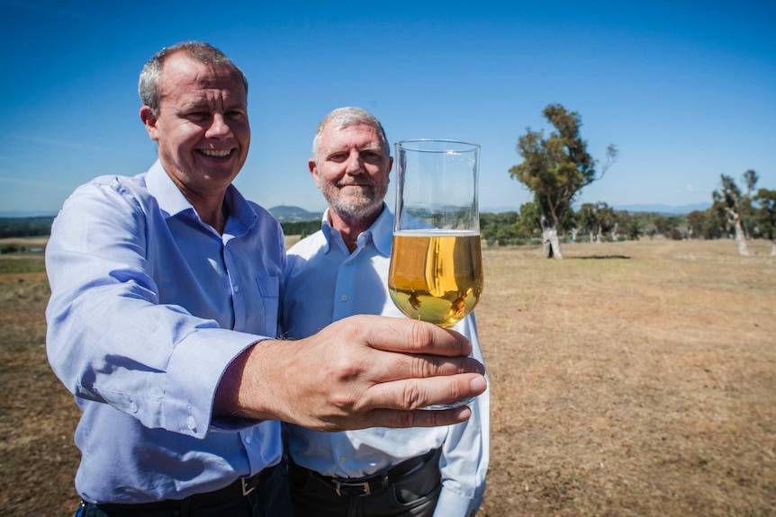 Two men standing in a paddock holding a glass of beer.