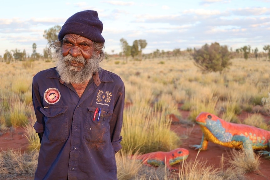 An Aboriginal man wearing a black beanie and blue shirt looks at the camera in front of a sculpture of the Great  Desert Skink.