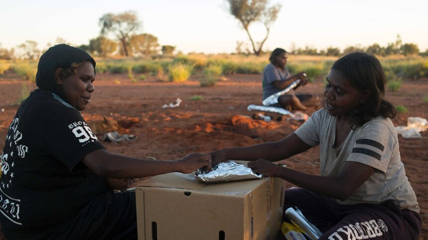 Three Indigenous women sit on red earth wrapping meat in foil. Two women are leaning on a cardboard box