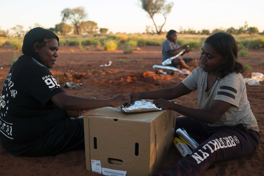 Three Indigenous women sit on red earth wrapping meat in foil. Two women are leaning on a cardboard box