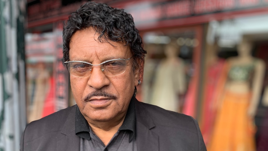 A man in a shirt and jacket stands outside a shop displaying saris in the window.