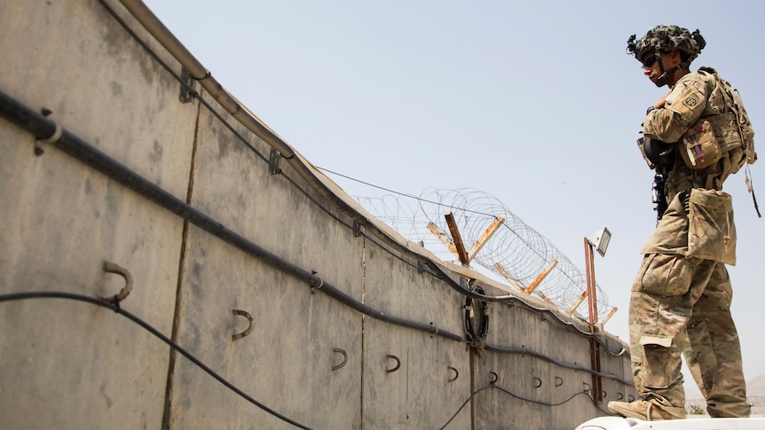 A soldier stands on the roof of a ute, parked next to a wall with razor wire on top. 