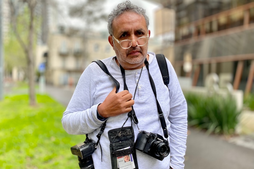 A middle-aged man with grey hair and stubble, wearing glasses, stands with two cameras draped over him and a a id card. 