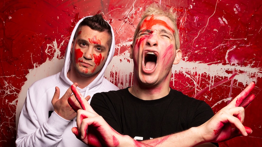Two brothers stand gesturing with their hands while covered in red paint