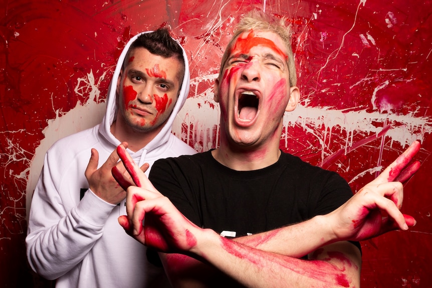 Two brothers stand gesturing with their hands while covered in red paint