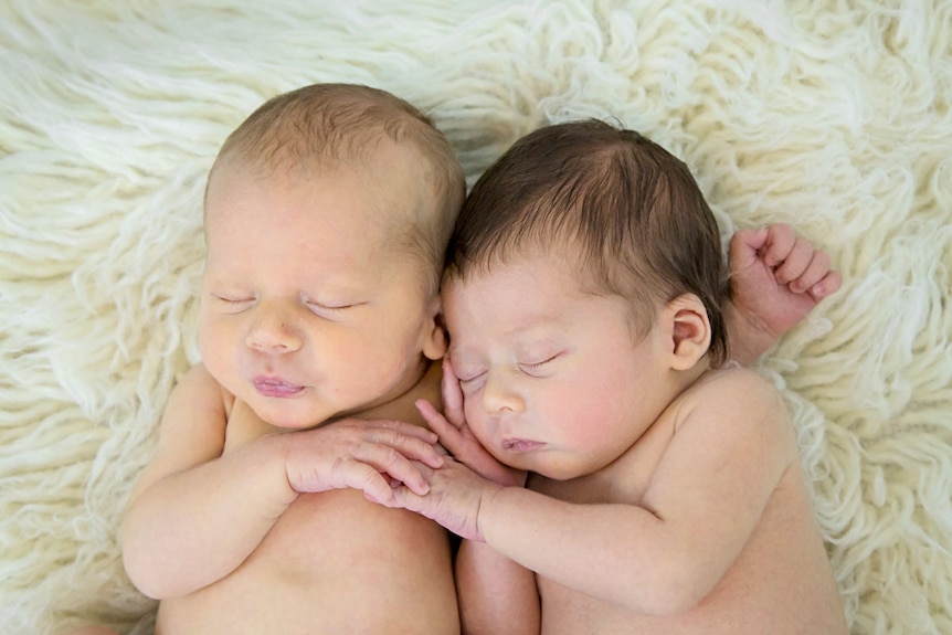 Two newborn babies cuddle together on a rug.