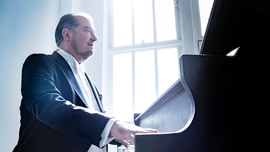 American pianist Garrick Ohlsson, wearing tails, plays a grand piano in front of a white sunlit window pane