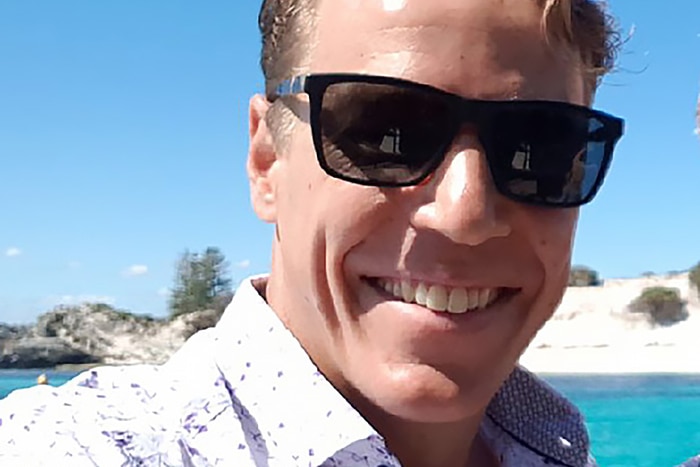 A tight head and shoulders shot of a man smiling and wearing sunglasses with a beach behind him.