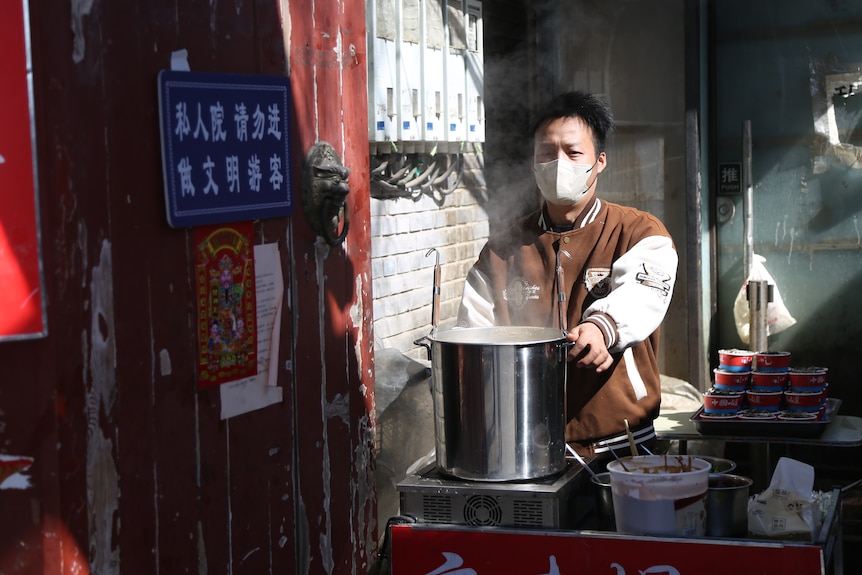 A young man wearing a mask holds a large stainless steel pot in a well-lit food shop.