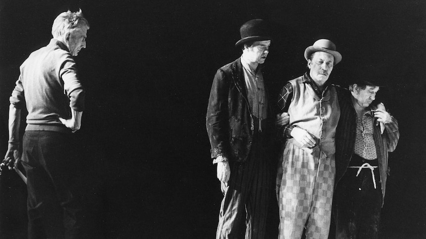 Samuel Beckett (left) observes three actors in hats on a stage.