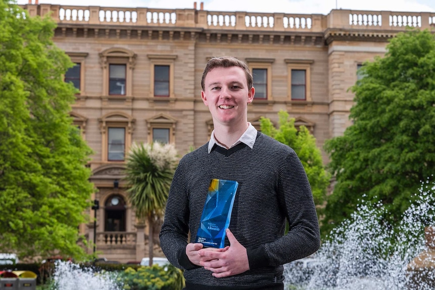 A young man stands with a glass trophy in front of a fountain and a sandstone building.