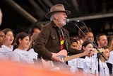 A white man with a brown hat and jacket holding a guitar and singing into a microphone surrounded by a group of kids.