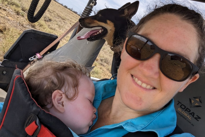 A woman in a blue shirt, with sunglasses holds her sleeping baby with her pet kelpie standing behind her.