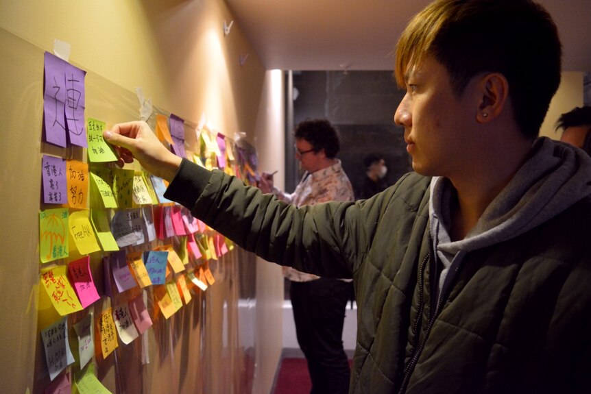 A man with tinted brown hair places a lime post it note with Chinese characters written on it.