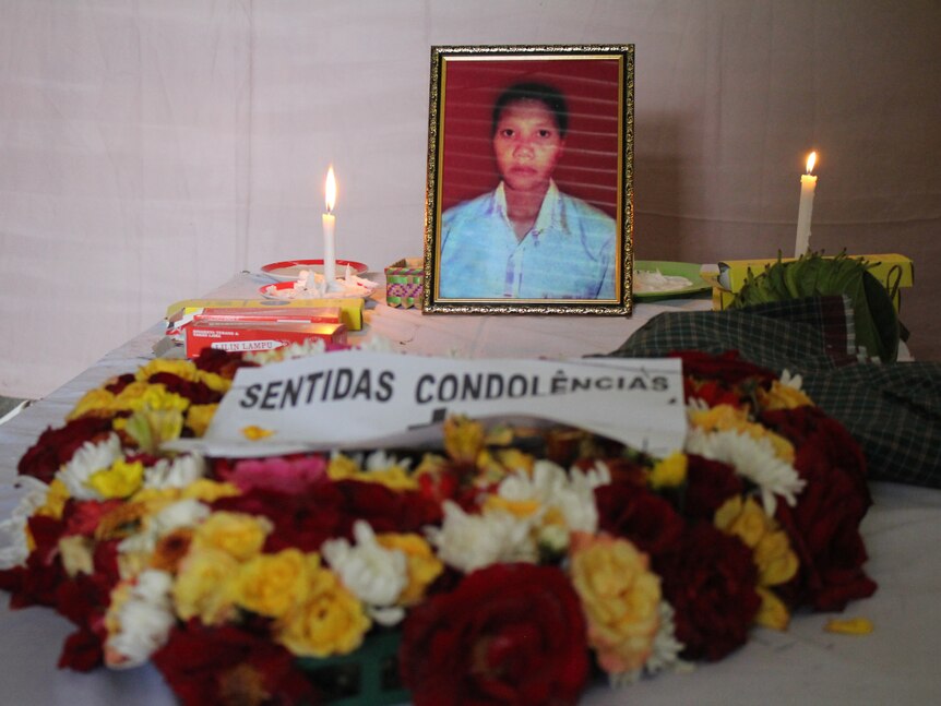 You view a close up of a woman's photo lying on a white coffin with flowers and candles placed over its top.