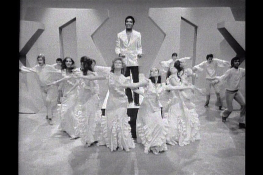 Black and white photo of Kamahl singing surrounded by dancers in TV studio.