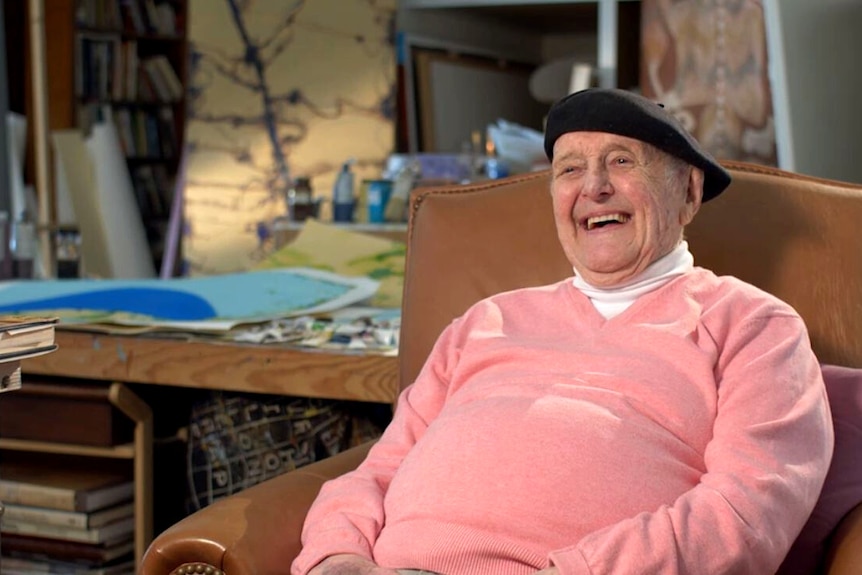 An elderly man in a beret and pink sweater sits in a chair in a busy art studio, smiling