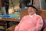 An elderly man in a beret and pink sweater sits in a chair in a busy art studio, smiling