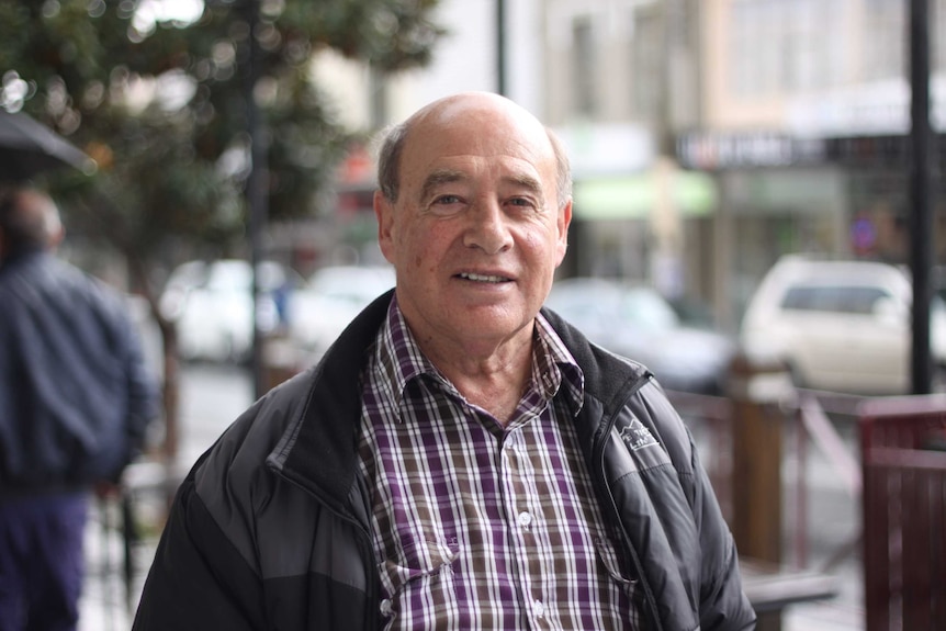 A man approx. 60 years of age, wearing a checked, button down shirt and puffy winter jacket standing on a main street.