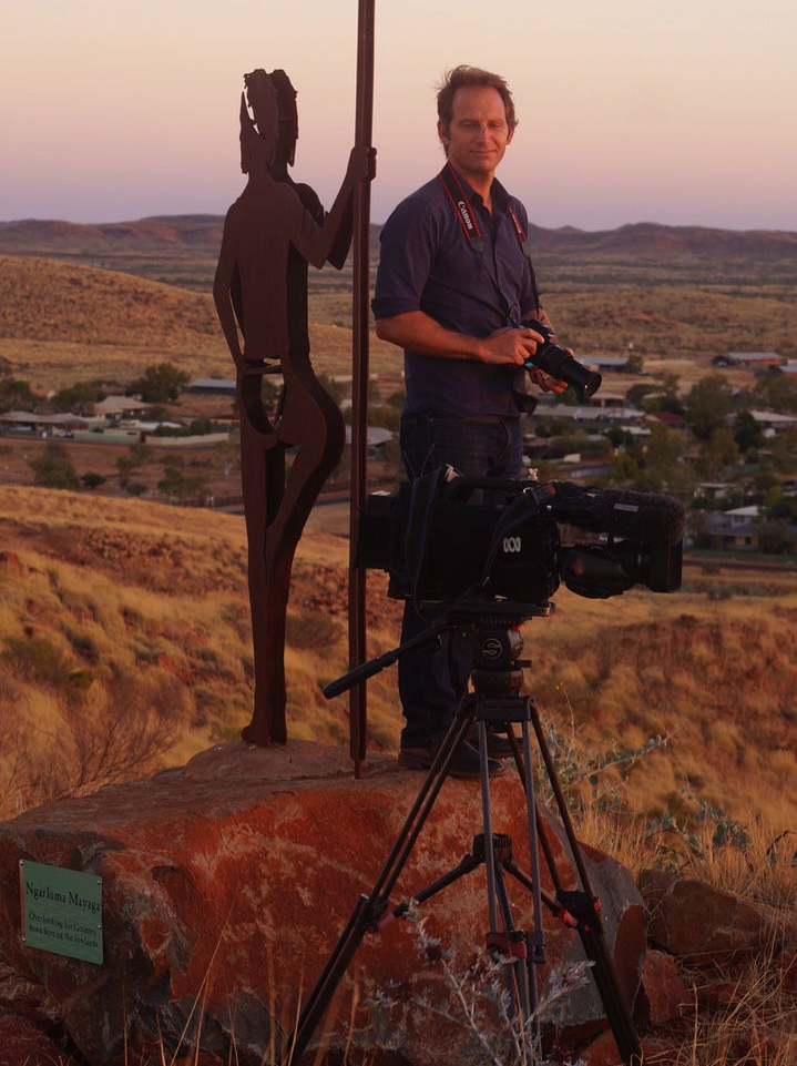 Journalist Nicolas Perpitch holding stills camera and standing next to indigenous statue in WA town of Roebourne.