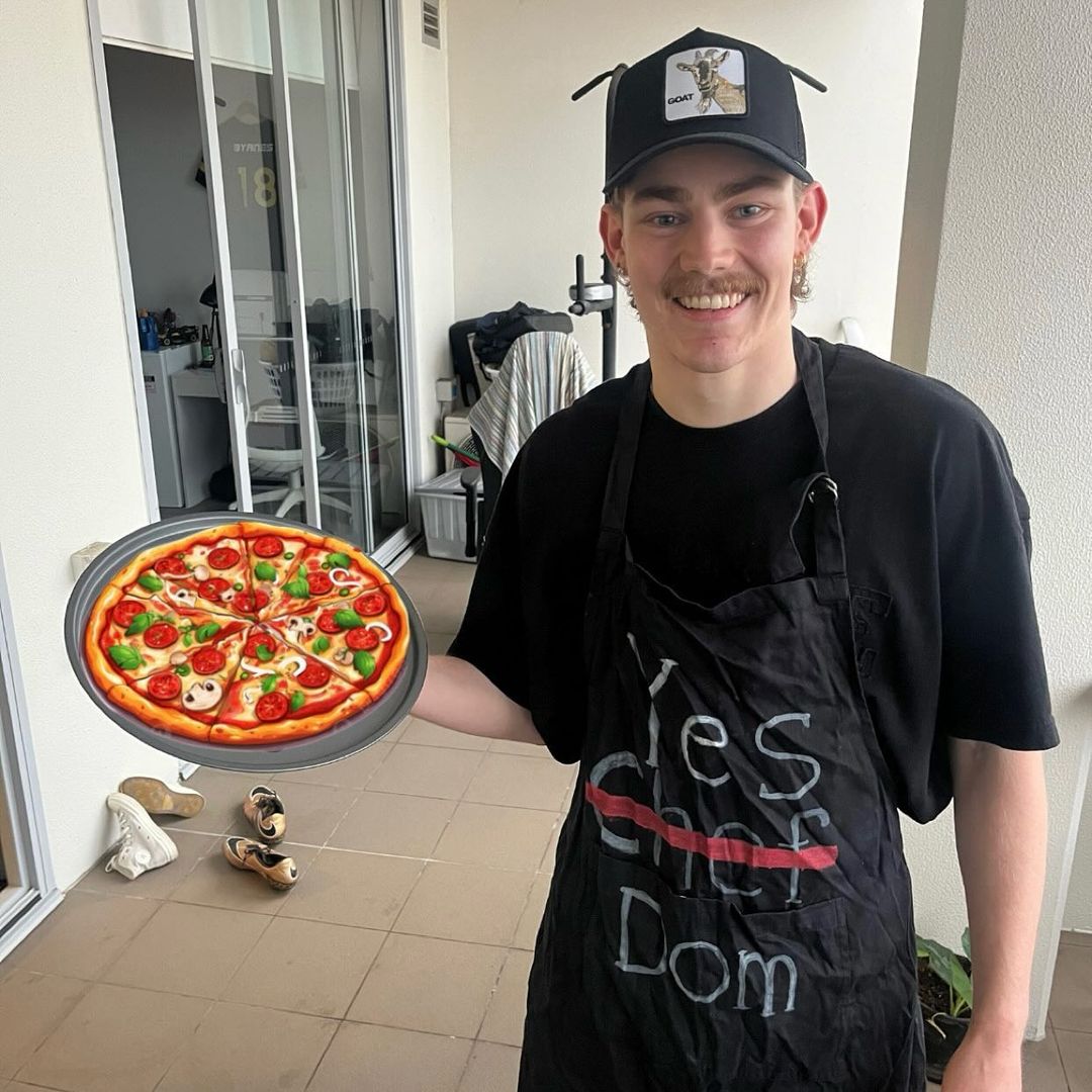 A man smiling at the camera with an apron on and holding a pizza