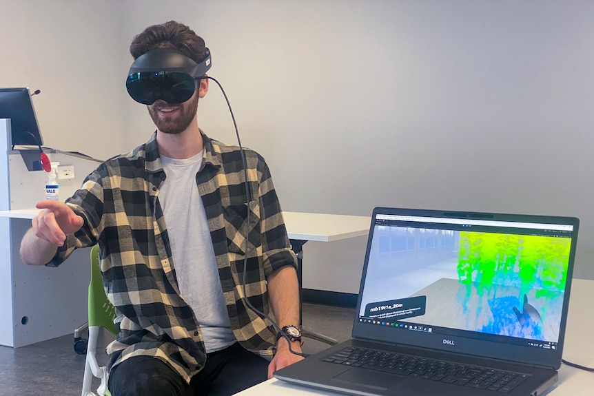 A man sits on a chair wearing a VR headset next to a laptop. 