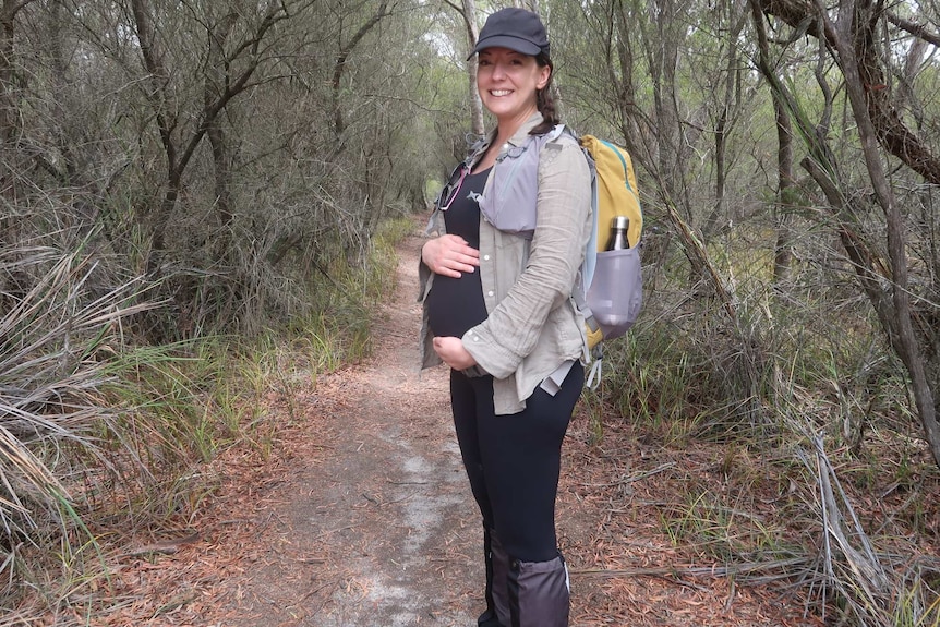Rachel Lees, pregnant, holding tummy and wearing backpack, with bush in background.