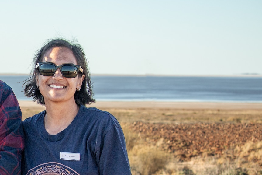 A woman in a navy shirt saying 'ARABANA' and sunglasses stands smiling in front of Kati Thanda-Lake Eyre. 