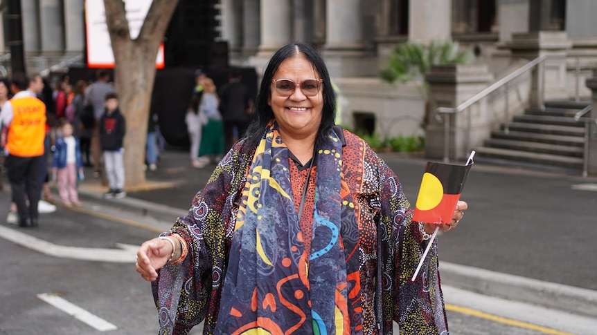 A smiling woman holds an Aboriginal flag in one hand standing in front of the steps of parliament house