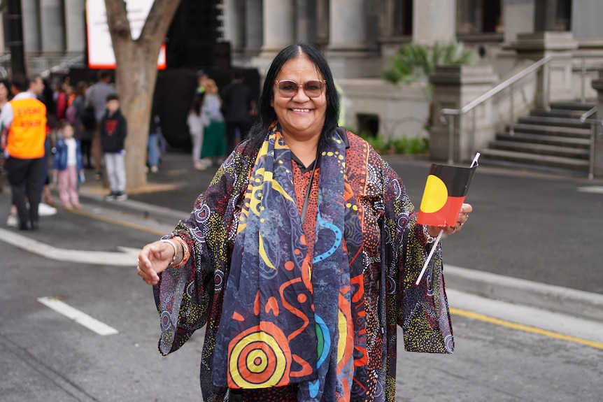 A smiling woman holds an Aboriginal flag in one hand standing in front of the steps of parliament house
