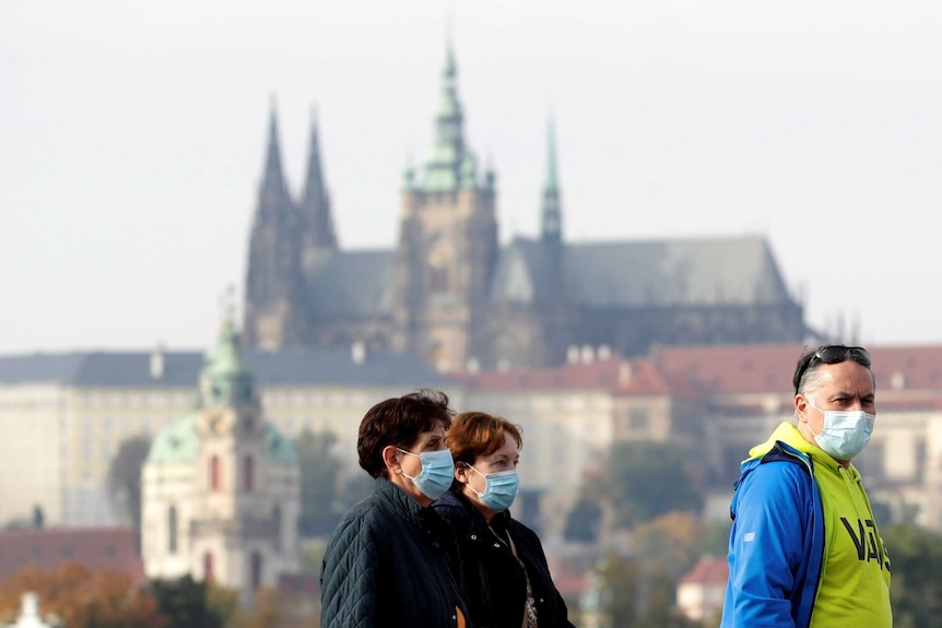 People wearing masks walk in street, a castle is in the background.