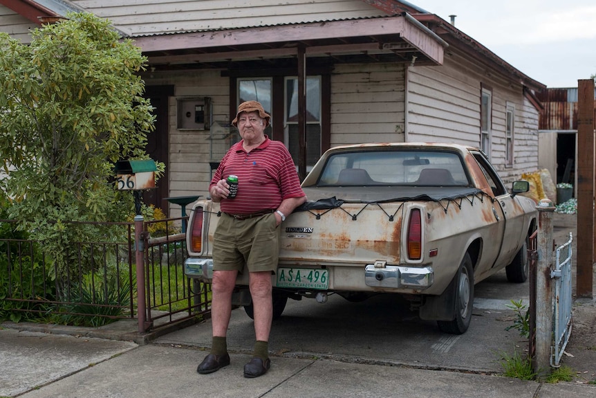 A man stands, holding a can of beer, next to a rusted ute outside his weatherboard house.