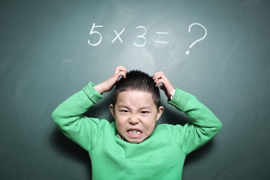 A boy with a frustrated expression in front of a blackboard showing a maths equation.