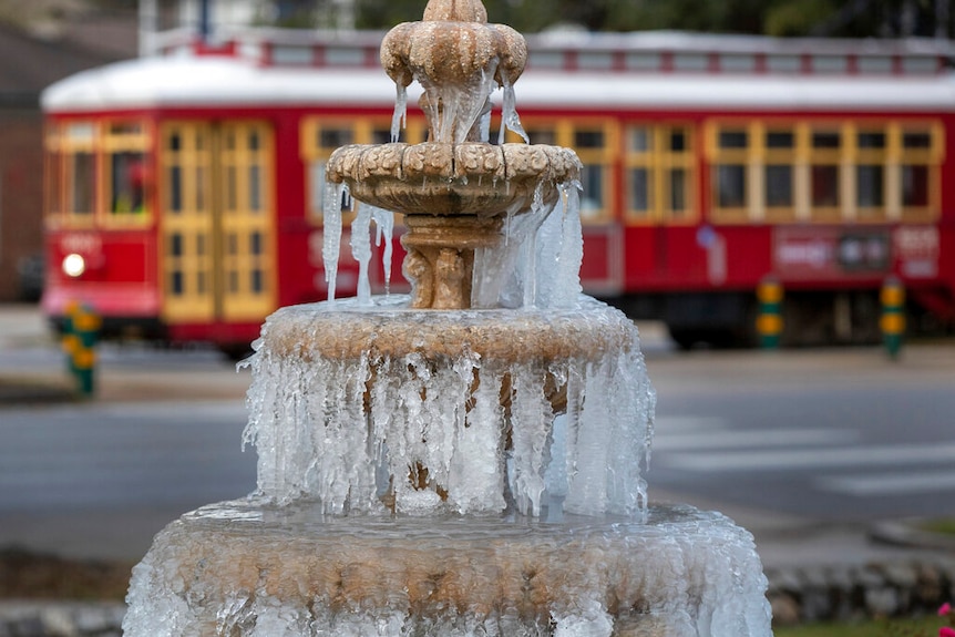 A fountain is seen frozen, icicles dripping down as a tram rumbles past in the background.