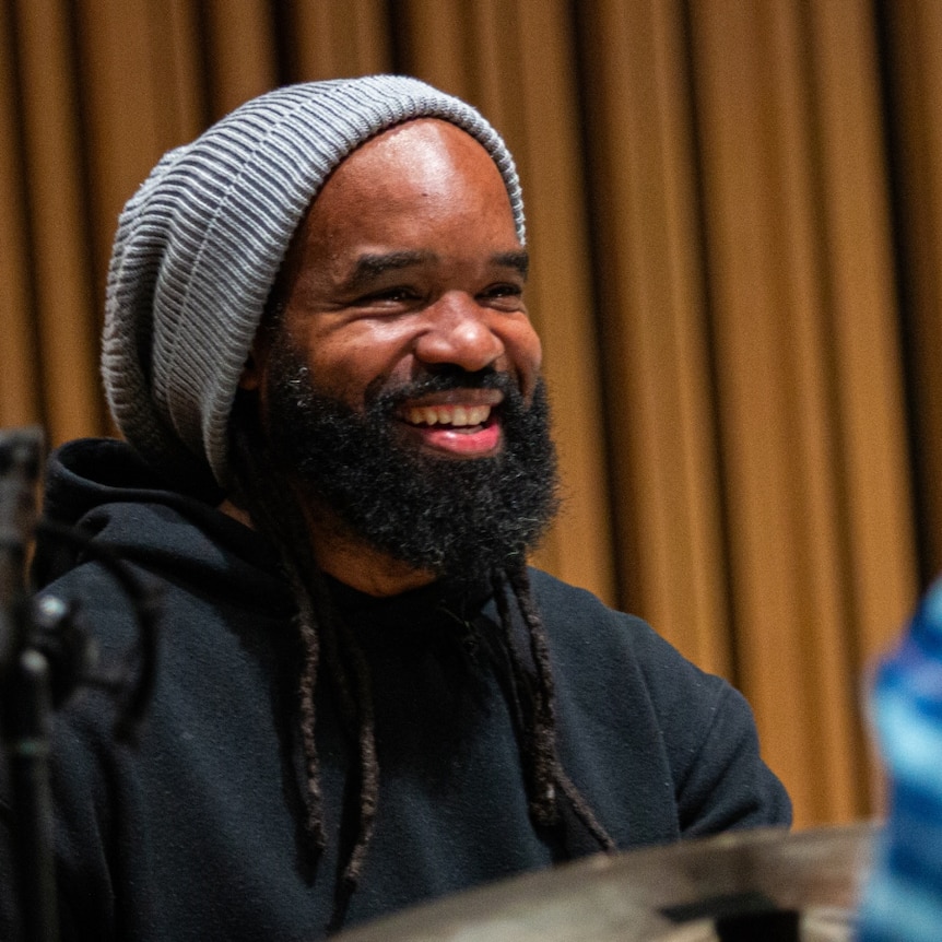 A colour photo of Obed Calvaire at the drums; he's wearing a beanie and smiling