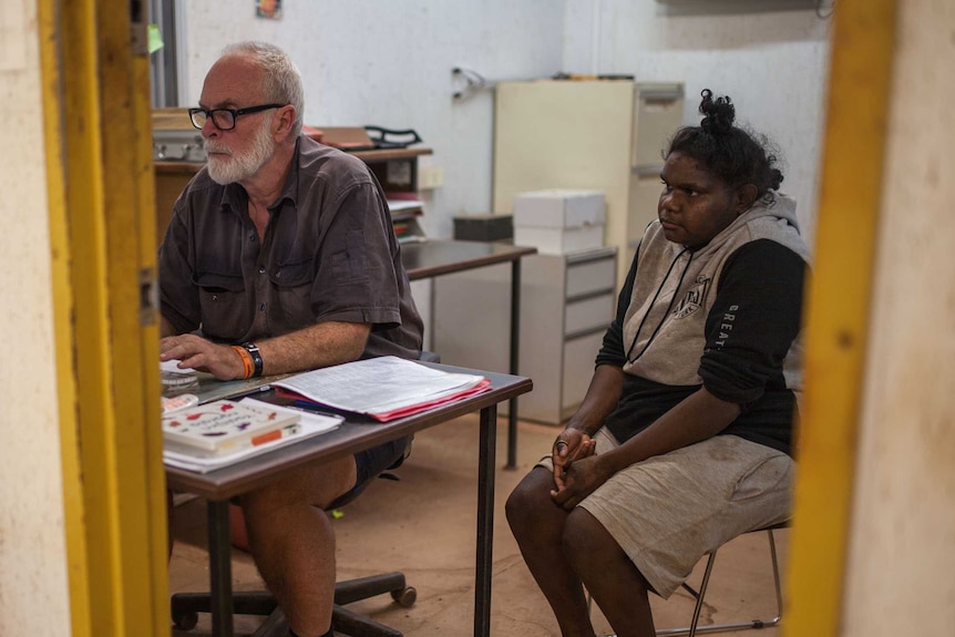 An older man with a white beard operates a computer while a young woman watches on.