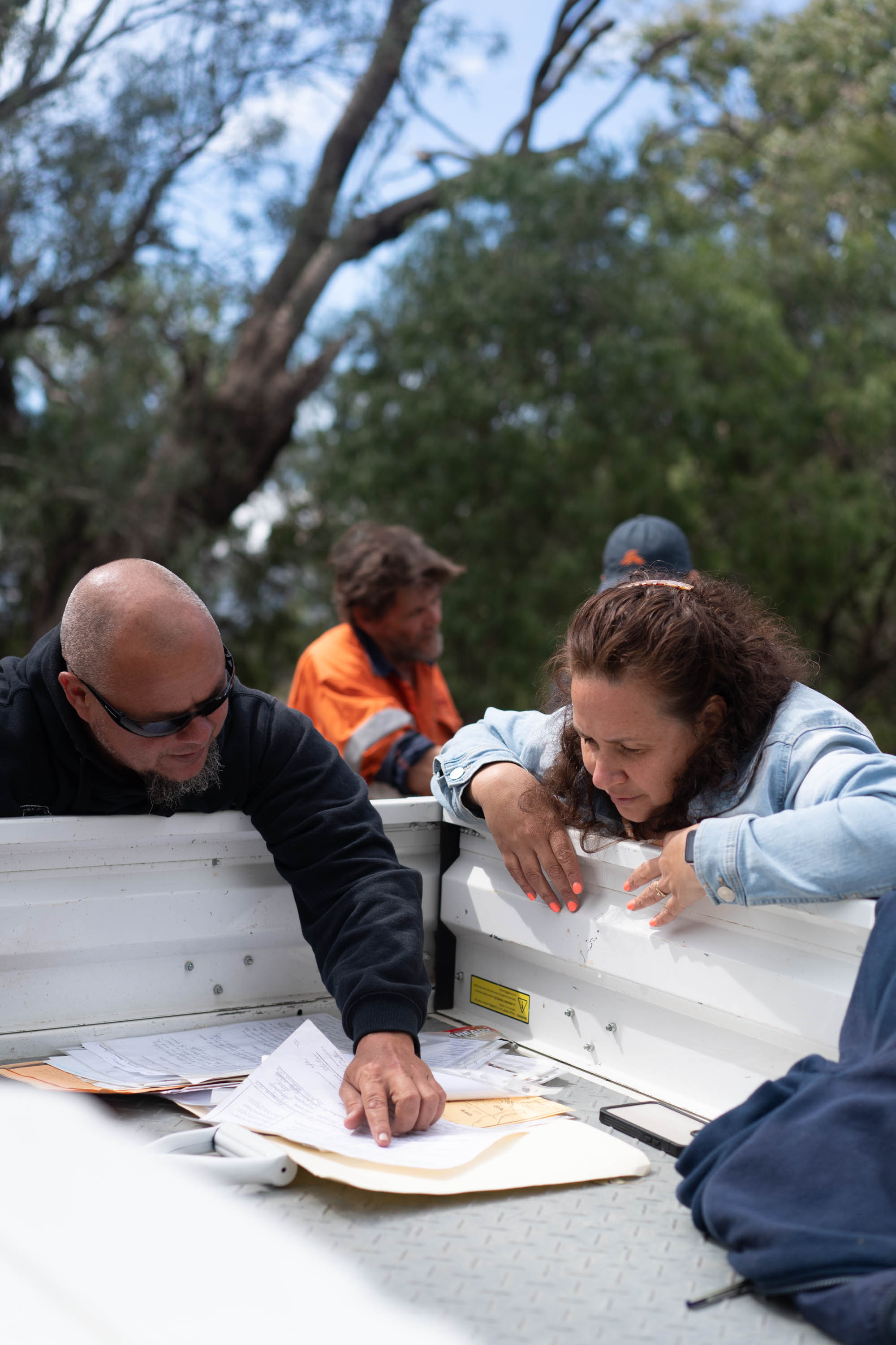 Man with shaved head and woman with long dark hair stand at back of ute back tray looking down at document on sunny day.