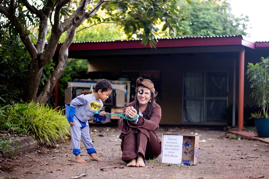 A young boy dressed as batman pictured with a woman dressed as pirate with a sign saying this too shall pass.