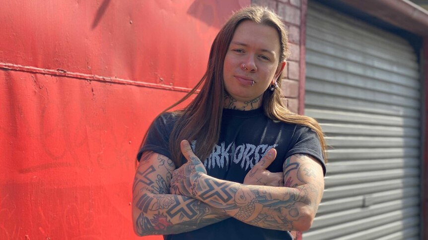 A person with long hair, a nose and lip ring, standing with tattooed arms crossed in front of a red wall and roller garage door.
