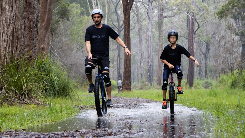 A man and his son ride unicycles along a wet bush track in the rain.