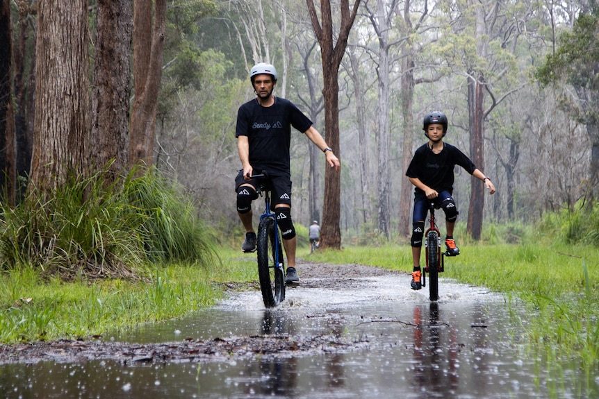 A man and his son ride unicycles along a wet bush track in the rain.