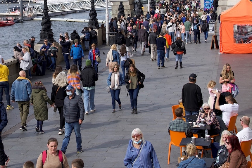 A crowd of people walk around and sit at tables next to the Thames