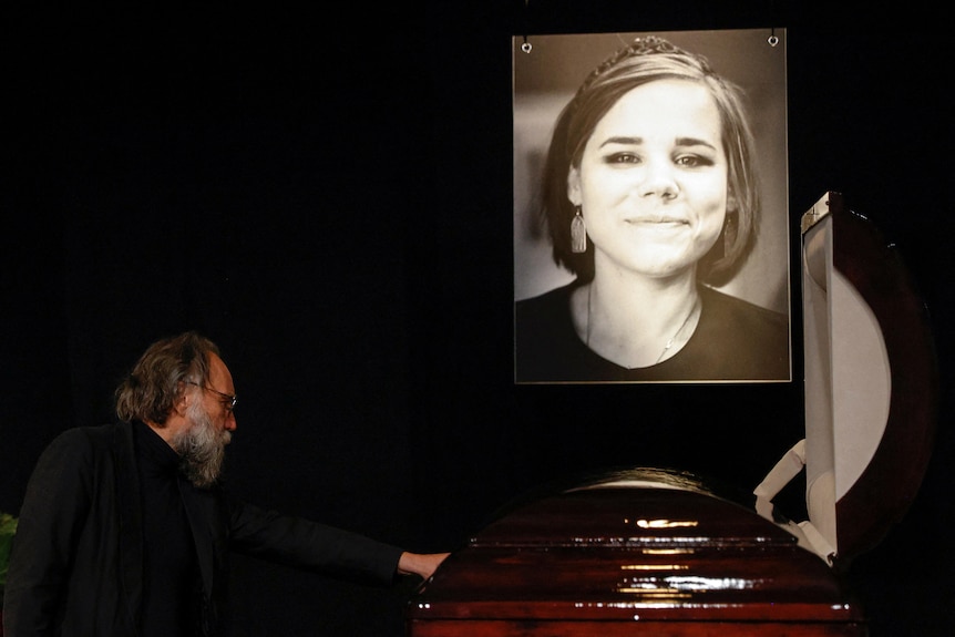A bearded man gently touches a coffin, with a young woman's photo hung above it