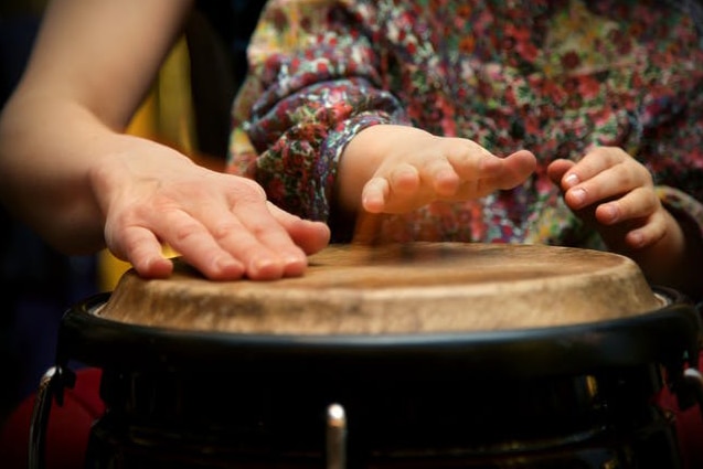 A child's hand plays a bongo-style drum next to an adult hand.