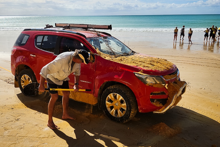 A man cleans the sand off one of the cars buried by the land slip a group of tourists look on from the water line on the beach