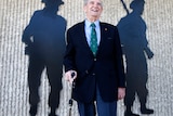 A man stands holding a walking stick in front of a wall that has two black cut outs of soldiers on it.