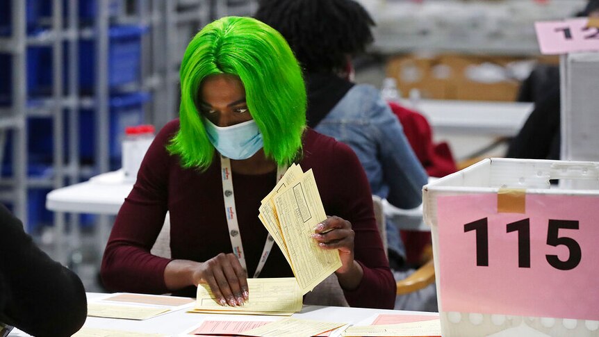 An election worker with bright green hair counts votes while wearing a face mask.