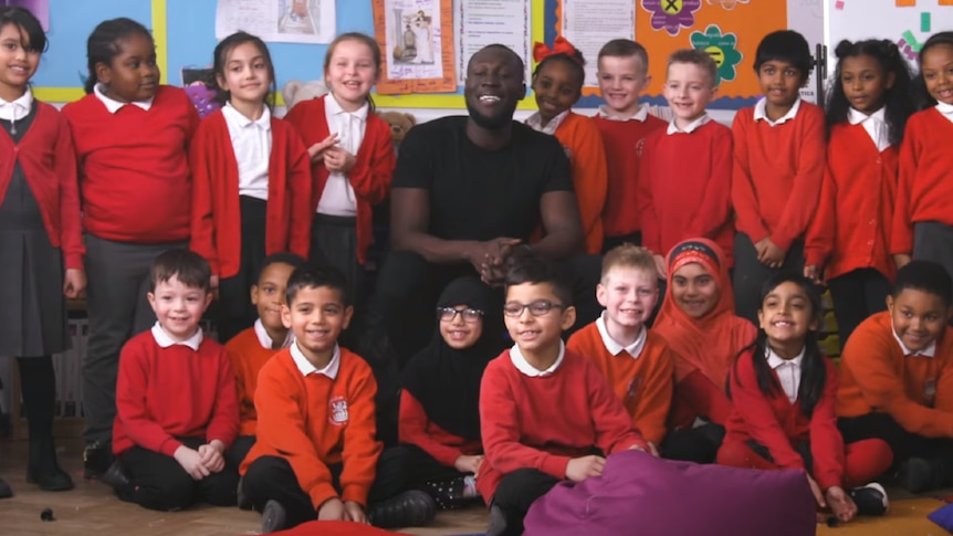 School children dressed in red, white and grey uniforms gathered around British rapper Stormzy, dressed in all black